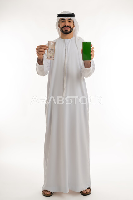 الأعمال الاقتصادية المالية، محاولة إدارة المصروف المنزلي بتدبر، بورتريه لرجل عربي خليجي اماراتي يرتدي الكندورة والغترة البيضاء ينظر الى الكاميرا بايماءات السعادة والسرور، عرض شاشة خضراء فارغة عبر الهاتف المحمول مع عملة الدرهم الاماراتي، خلفية بيضاء، كروما