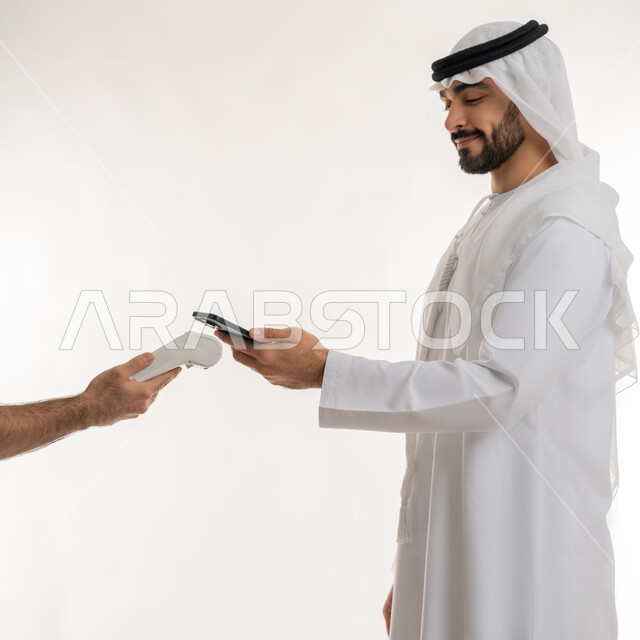 تسهيل عمليات الشراء والبيع، استخدام الوسائل والتقنيات الحديثة المتطورة، إمكانية تسديد الفواتير عن طريق جهاز الدفع الالكتروني، بورتريه من الجانب لرجل عربي خليجي اماراتي يرتدي الكندورة يسدد عند أمين الصندوق باستخدام جهاز الهاتف المحمول، خلفية بيضاء