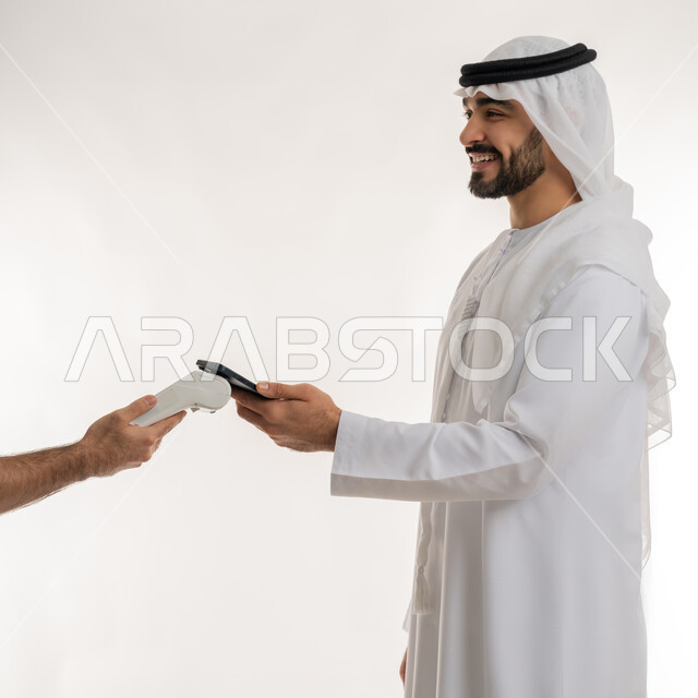 Facilitating buying and selling operations, using modern advanced means and technologies, the possibility of paying bills via an electronic payment device, a side portrait of an Arab Gulf Emirati man wearing a kandura paying at the cashier using a mobile phone, white background