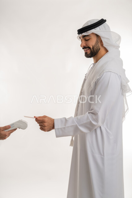 Using modern advanced means and technologies, the possibility of paying bills using electronic cards, a portrait from the side of an Arab Gulf Emirati man wearing a white kandura and ghutra paying using a bank credit card, facilitating buying and selling operations, white background