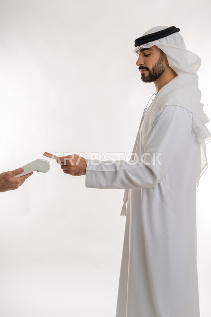 تسهيل عمليات الشراء والبيع، إمكانية تسديد الفواتيرعن طريق البطافات الالكترونية، استخدام الوسائل والتقنيات الحديثة المتطورة، بورتريه من الجانب لرجل عربي خليجي اماراتي يرتدي الكندورة والغترة البيضاء يقوم بالدفع باستخدام بطاقة الإئتمان البنكية، خلفية بيضاء