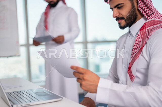 رجل اعمال عربي خليجي سعودي يجلس امام الحاسوب المحمول ، يحمل بيده ورقة المخطط المالي ، اجتماع فريق العمل ، مناقشة الرسم البياني المالي ، خطة الاستثمار ، استشارات مالية ومحاسبية، تحقيق اهداف العمل