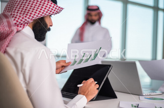 رجل اعمال عربي خليجي سعودي يجلس امام الحاسوب المحمول ، يحمل بيده ورقة المخطط المالي ، اجتماع فريق العمل ، مناقشة الرسم البياني المالي ، خطة الاستثمار ، استشارات مالية ومحاسبية، تحقيق اهداف العمل