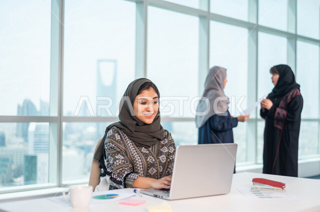 A Saudi Arabian Gulf employee sitting in front of a laptop, behind her are two Saudi Arabian female employees, discussing work-related issues, a work team within a Saudi company, completing work tasks, achieving company goals, developing work