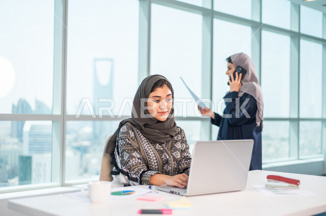 موظفة عربية خليجية سعودية تجلس امام الحاسوب المحمول ، خلفها موظفة عربية سعوية خليجية تتحدث عبر الهاتف المحمول ، فريق عمل داخل شركة سعودية ، انجاز مهام العمل ، تحقيق اهداف الشركة ، تطوير العمل  