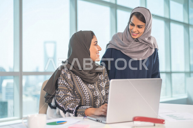 موظفة عربية خليجية سعودية تجلس امام الحاسوب المحمول ، بجانبها موظفة عربية سعوية خليجية تتناقشان امور خاصة بالعمل، فريق عمل داخل شركة سعودية ،متابعة البورصة ،استشارات مالية ومحاسبية ، تحقيق اهداف العمل