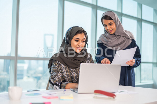 موظفة عربية خليجية سعودية تضع سماعة الراس و تجلس امام الحاسوب المحمول، بجانبها موظفة عربية سعوية خليجية تتناقشان امور خاصة بالعمل، فريق عمل داخل شركة سعودية ،متابعة البورصة ،استشارات مالية ومحاسبية ، تحقيق اهداف العمل