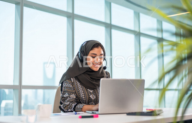موظفة عربية خليجية سعودية تضع سماعة الراس و تجلس امام الحاسوب المحمول ، اجراء اتصال فيديو من خلال الجهاز المحمول ، عقد اجتماع عمل عبر الانترنت ، العمل عن بعد ، متابعة وتطوير العمل