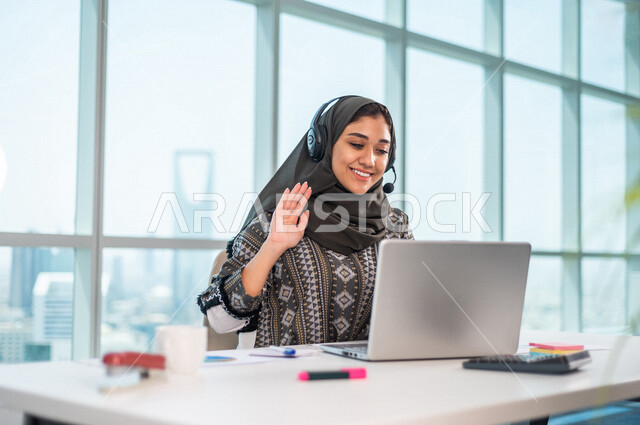 موظفة عربية خليجية سعودية تضع سماعة الراس و تجلس امام الحاسوب المحمول ، اجراء اتصال فيديو من خلال الجهاز المحمول ، عقد اجتماع عمل عبر الانترنت ، العمل عن بعد ، متابعة وتطوير العمل