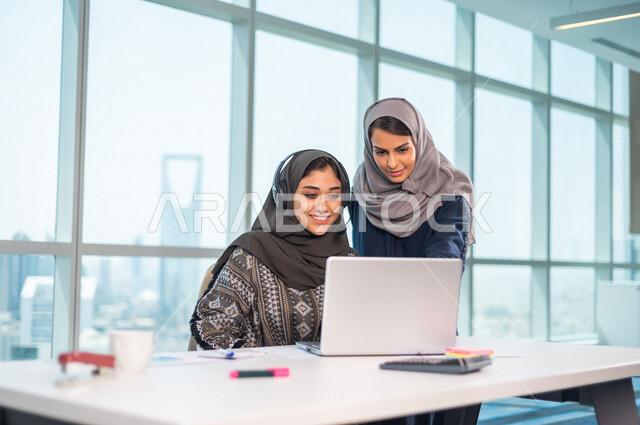 موظفة عربية خليجية سعودية تضع سماعة الراس و تجلس امام الحاسوب المحمول، بجانبها موظفة عربية سعوية خليجية تتناقشان امور خاصة بالعمل، فريق عمل داخل شركة سعودية ،متابعة البورصة ،استشارات مالية ومحاسبية ، تحقيق اهداف العمل