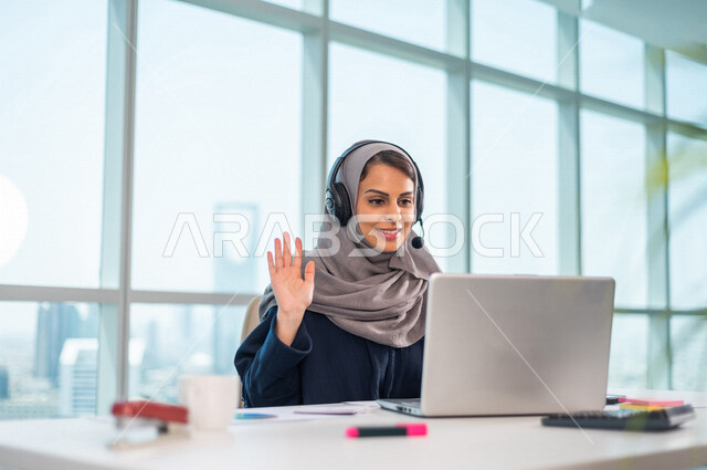 موظفة عربية خليجية سعودية تضع سماعة الراس و تجلس امام الحاسوب المحمول ، اجراء اتصال فيديو من خلال الجهاز المحمول ، عقد اجتماع عمل عبر الانترنت ، العمل عن بعد ، متابعة وتطوير العمل