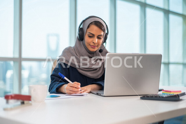 A Saudi Arabian Gulf employee puts on a headset and sits in front of a laptop, making a video call through a mobile device, holding a work meeting via the Internet, taking notes on paper, working remotely, following up and developing work