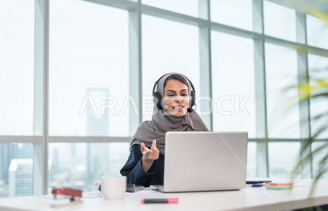 موظفة عربية خليجية سعودية تضع سماعة الراس و تجلس امام الحاسوب المحمول ، اجراء اتصال فيديو من خلال الجهاز المحمول ، عقد اجتماع عمل عبر الانترنت ، العمل عن بعد ، متابعة وتطوير العمل