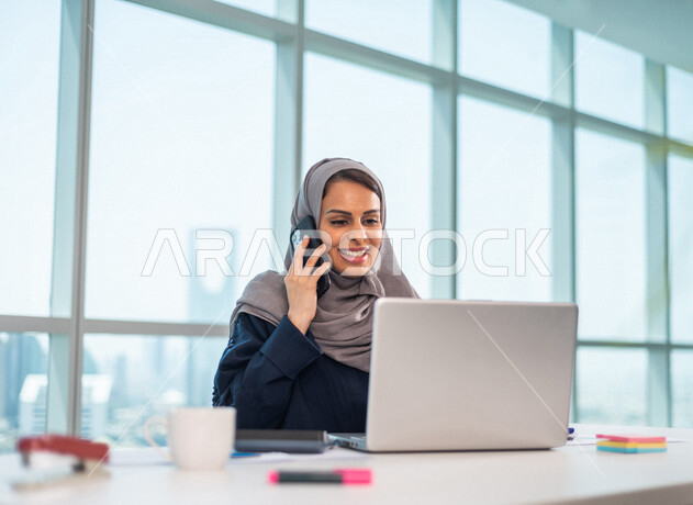 موظفة عربية خليجية سعودية تجلس امام الحاسوب المحمول ، اجراء اتصال من خلال الهاتف المحمول ، مناقشة امور خاصة بالعمل ،  انجاز مهام العمل ، تحقيق اهداف الشركة ، تطوير العمل  