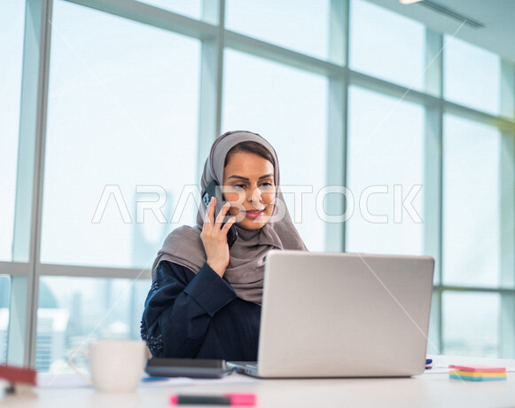 موظفة عربية خليجية سعودية تجلس امام الحاسوب المحمول ، اجراء اتصال من خلال الهاتف المحمول ، مناقشة امور خاصة بالعمل ،  انجاز مهام العمل ، تحقيق اهداف الشركة ، تطوير العمل  