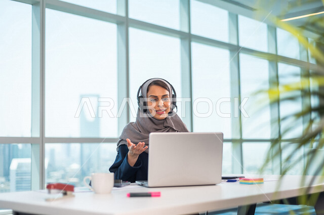 موظفة عربية خليجية سعودية تضع سماعة الراس و تجلس امام الحاسوب المحمول ، اجراء اتصال فيديو من خلال الجهاز المحمول ، عقد اجتماع عمل عبر الانترنت ، العمل عن بعد ، متابعة وتطوير العمل