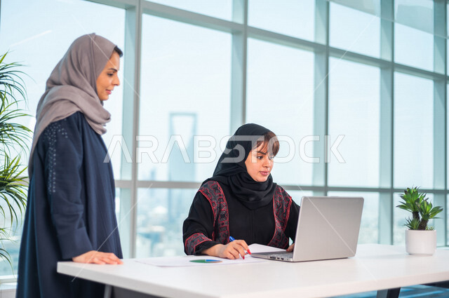 موظفة عربية خليجية سعودية تجلس امام الحاسوب المحمول ، بجانبها موظفة عربية سعوية خليجية تتناقشان امور خاصة بالعمل، فريق عمل داخل شركة سعودية ،متابعة البورصة ،استشارات مالية ومحاسبية ، تحقيق اهداف العمل