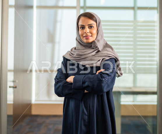 موظفة عربية خليجية سعودية تقف مبتسمة وسعيدة امام الكاميرا، خلفها المكتب الخاص بالعمل ، سيدة اعمال تعمل في شركة سعودية ، وظائف ومهن مكتبية