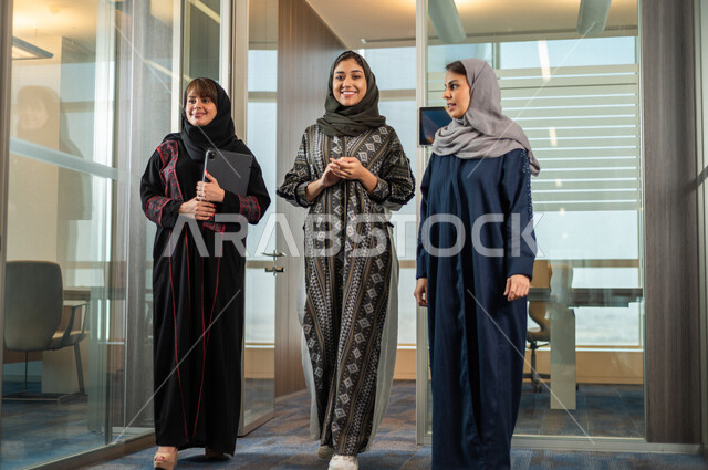 A group of Saudi Arabian Gulf female employees walking inside a Saudi company, a Gulf businesswoman holding a tablet device in her hand, completing work tasks, achieving company goals, developing work