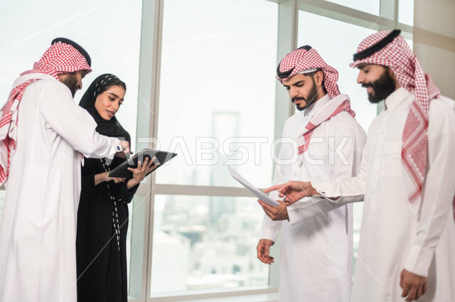 A group of Saudi businessmen inside the company's headquarters, discussing the agenda and tasks, an Arab Saudi Gulf secretary working on organizing tasks and work and taking notes through a technical device, unity and teamwork