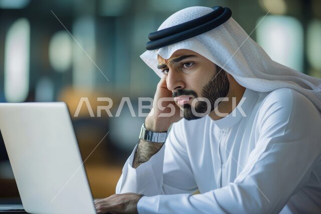 Using modern and advanced technical devices to accomplish tasks, a young Saudi Arabian Gulf man wearing the traditional thobe and shemagh works on a laptop, putting his hand on his cheek with an expression of boredom and displeasure