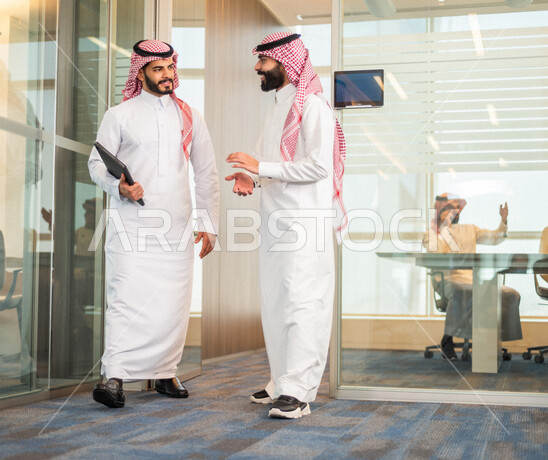 موظف عربي خليجي سعودي يحمل بيده جهاز التابلت ، موظفان عربيان خليجيان يتناقشان امور خاصة بالعمل ، الوحدة والعمل الجماعي ، تحقيق اهداف الشركة، تطوير العمل