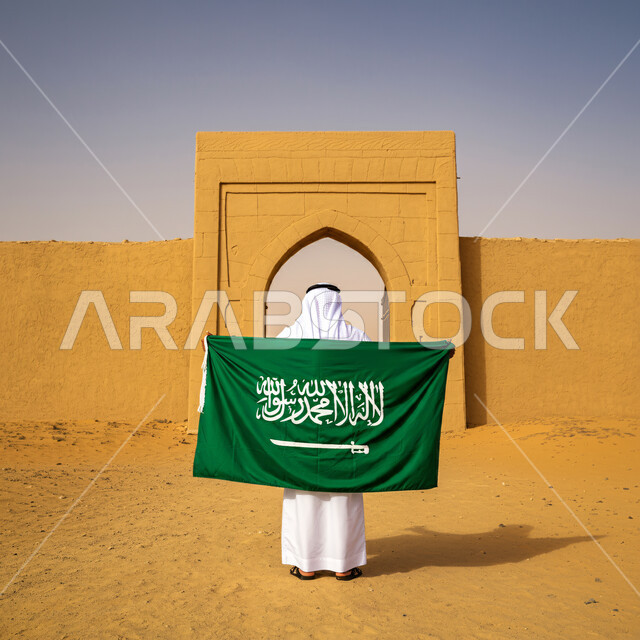 Pride and pride in national identity, a back view of a Saudi Arabian Gulf man wearing traditional thobe and shemagh standing straight holding the flag of the Kingdom of Saudi Arabia, Saudi National Day commemoration March 23, Saudi Flag Day March 11, love of the homeland and belonging to it