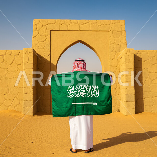 Pride and pride in national identity, a back view of a Saudi Arabian Gulf man wearing traditional thobe and shemagh standing straight holding the flag of the Kingdom of Saudi Arabia, Saudi National Day commemoration March 23, Saudi Flag Day March 11, love of the homeland and belonging to it