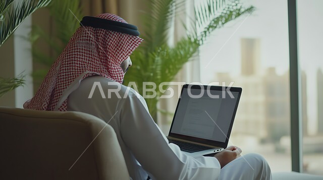مهن ووظائف ادارية مكتبية، دمج العمل مع التكنولوجيا، مفهوم إدارة شؤون الأعمال وتنظيمها عبر الاجهزة التقنية، صورة مقربة من الخلف لرجل اعمال عربي خليجي سعودي يرتدي الزي التقليدي يقوم بالعمل على الحاسوب داخل المكتب