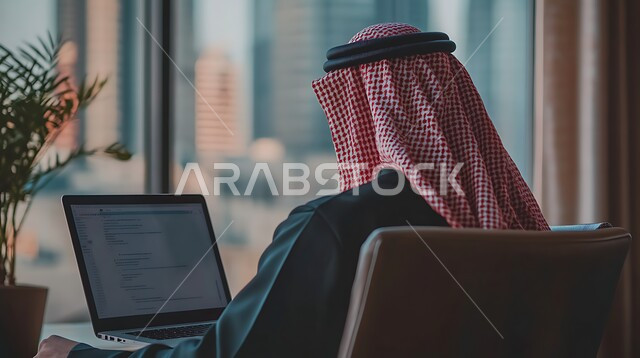 Integrating work with technology, office administrative professions and jobs, the concept of managing and organizing business affairs through technical devices, a close-up image from the back of a Saudi Arabian Gulf businessman wearing traditional clothing working on the computer inside the office