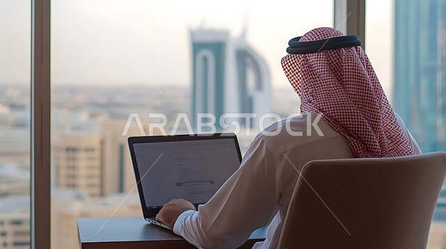 مفهوم إدارة شؤون الأعمال وتنظيمها عبر الاجهزة التقنية، دمج العمل مع التكنولوجيا، مهن ووظائف ادارية مكتبية، صورة مقربة من الخلف لرجل اعمال عربي خليجي سعودي يرتدي الزي التقليدي يقوم بالعمل على الحاسوب داخل المكتب