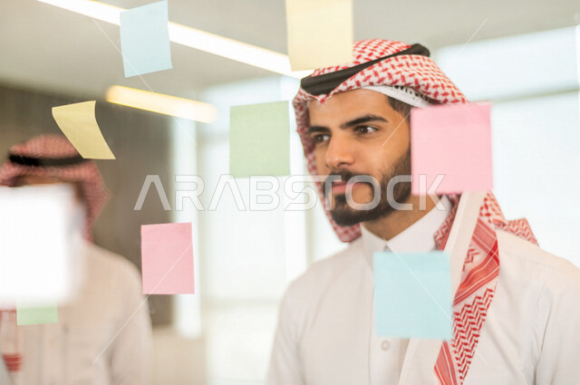 موظفان عربيان خليجيان سعوديان ينظران الى الملاحظات المعلقة ، رجال اعمال خليجيين سعوديين يتشاركون بتحديد الاهداف ، انجاز مهام العمل ، تحقيق اهداف الشركة ، تطوير العمل