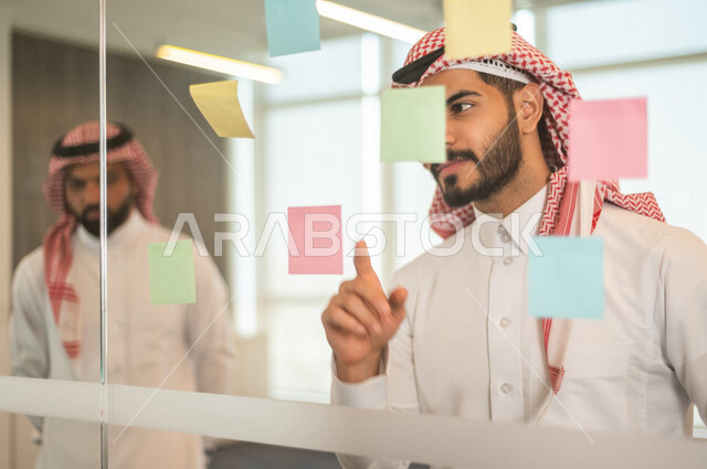 موظفان عربيان خليجيان سعوديان ينظران الى الملاحظات المعلقة ، رجال اعمال خليجيين سعوديين يتشاركون بتحديد الاهداف ، انجاز مهام العمل ، تحقيق اهداف الشركة ، تطوير العمل