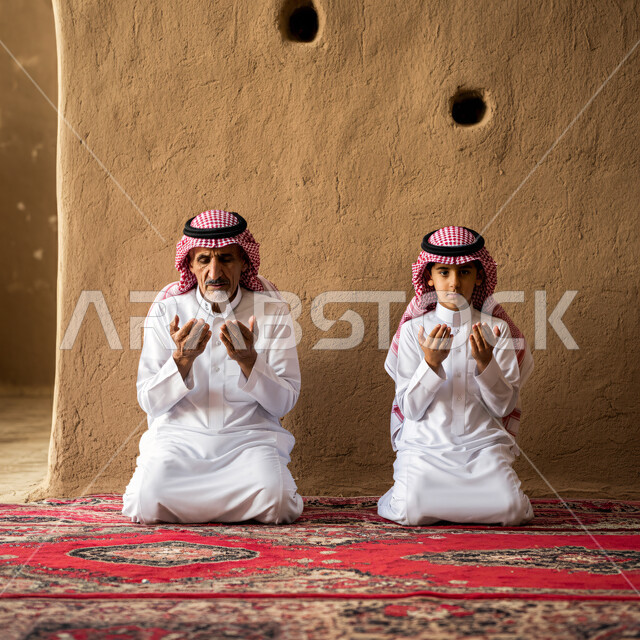 الإكثار من الطاعات وذكر الله تعالى، رجل كبير بالسن عربي سعودي خليجي يجلس على السجادة يؤدي فرض الصلاة بجانب ابنه الصبي يرفعان أيديهما يقومان بالدعاء، التقرب إلى رب العالمين في شهر رمضان، الاجتهاد في العبادة وأداء الفرائض الدينية، أجواء عائلية رمضانية روحانية