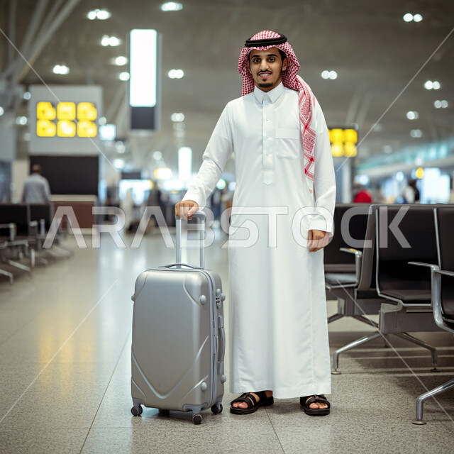 الاستعداد للذهاب في رحلة ترفيهية، رجل عربي خليجي سعودي يرتدي الشماغ والثوب التقليدي يمسك بيده حقيبة سفر ينظر إلى الكاميرا بإيماءات السعادة والسرور، صالة الانتظار في مطارات المملكة العربية السعودية، قضاء إجازة صيفية ممتعة