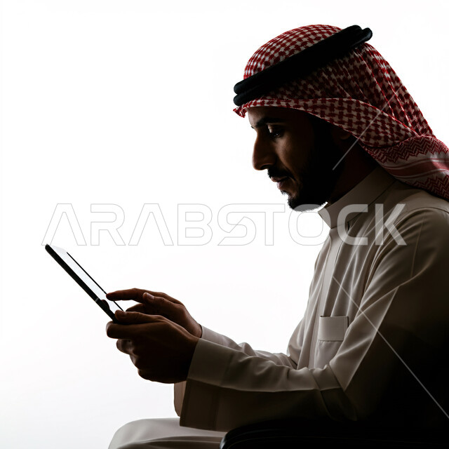Chatting with friends via tablet programs and applications, browsing social networking sites, close-up side portrait of a Saudi Arabian Gulf man wearing a traditional shemagh and thobe sitting on a chair holding a tablet in his hand, using modern technologies, holding a business meeting online, white background