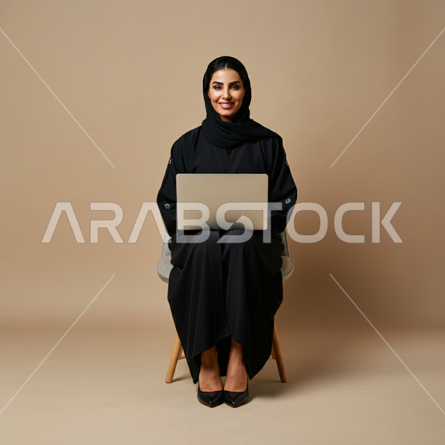 إنجاز مهام العمل عن بعد من خلال الإنترنت، بورتريه لامرأة عربية خليجية سعودية ترتدي الحجاب والعباءة تجلس على الكرسي تعمل على الحاسب المحمول تنظر إلى الكاميرا بإيماءات السرور، مهن ووظائف نسائية مكتبية، استخدام التكنولوجيا الحديثة والمتطورة، خلفية بيج