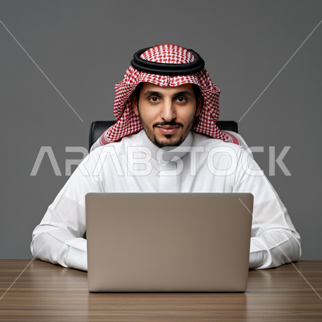 إدارة شؤون الأعمال وتنظيمها عبر الأجهزة الحديثة، مهن ووظائف إدارية مكتبية، دمج العمل مع التقنية والتكنولوجيا، بورتريه مقرب لرجل أعمال عربي خليجي سعودي يرتدي الزي التقليدي يعمل على الحاسوب المحمول داخل المكتب، متابعة ومناقشة الخطط وتحقيق أهداف الشركة، خلفية رمادية