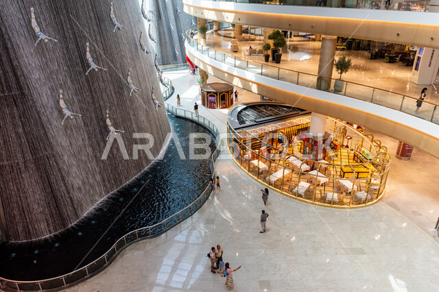  كشك لبيع العطور الفخمة، صورة من الأعلى لتماثيل فنية لغواصي اللؤلؤ وسط شلال داخل دبي مول، المعالم السياحية الترفيهية المشهورة في مدينة دبي، السياحة في الإمارات العربية المتحدة،  وجهات عائلية للتسوق، جذب واستقطاب السائحين من أنحاء العالم