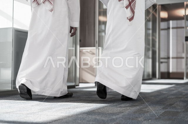 صورة من الخلف لرجلان اعمال عربيان خليجيان سعوديان يرتديان الزي التقليدي السعودي ، يمشيان في مقر الشركة ، يتناقشان في أمور خاصة بالعمل ، تحقيق أهداف الشركة و تطوير العمل ، متابعة استراتيجية العمل و المشروع ، شركة سعودية