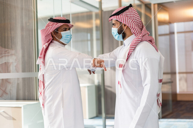 صورة لرجلان اعمال عربيان خليجيان سعوديان يرتديان الزي التقليدي السعودي و الكمامة للوقاية من فايروس كورونا  يتصافحان عن طريق الكوع في مقر الشركة ، ايماءات وتعابير لغة الجسد ، اتباع اجراءات الوقاية و الحماية من فايروس كورونا ، شركة سعودية
