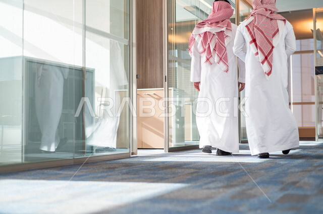 صورة من الخلف لرجلان اعمال عربيان خليجيان سعوديان يرتديان الزي التقليدي السعودي ، يمشيان في مقر الشركة ، يتناقشان في أمور خاصة بالعمل ، تحقيق أهداف الشركة و تطوير العمل ، متابعة استراتيجية العمل و المشروع ، شركة سعودية