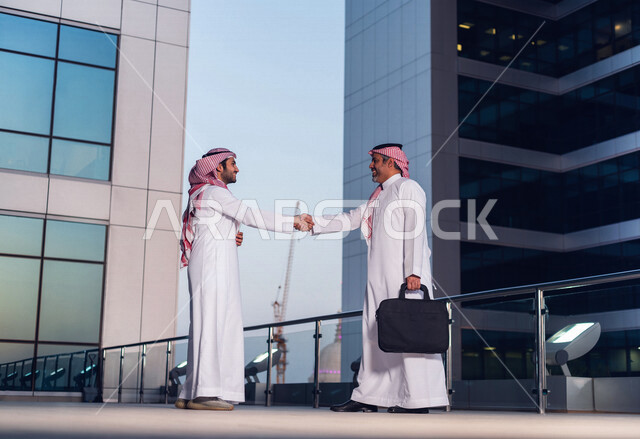 رجلان أعمال عربيان خليجيان سعوديان يرتديان الزي السعودي التقليدي يتصافحان داخل شركة سعودية ، اتفاق عمل ، عقد عمل جديد ، شراكة عمل