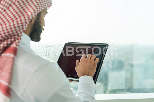 صورة لرجل اعمال عربي خليجي سعودي يرتدي الزي التقليدي السعودي ، يعمل في مقر الشركة ، يستخدم جهاز تقني لانجاز مهامه ، اشراف و متابعة ، متابعة استراتيجية عمل و مشروع ،  بيئة عمل ، شركة سعودية