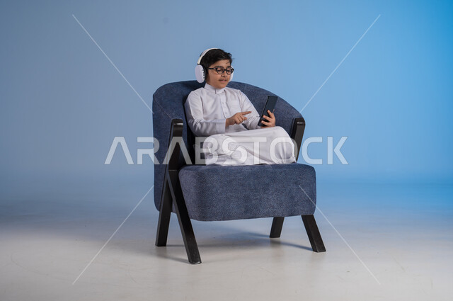 استخدام تقنية الهيدفون الحديثة, قضاء وقت ممتع بالاستماع إلى المقاطع الموسيقية المفضلة, بورتريه لفتى عربي خليجي سعودي يرتدي الثوب التقليدي يجلس على كنبه مريحة يضع سماعة الرأس الصوتية ويستخدم الهاتف المحمول, خلفية زرقاء