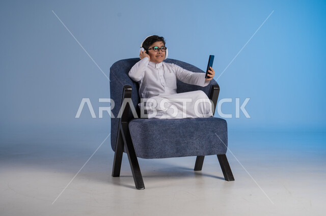 استخدام تقنية الهيدفون الحديثة, قضاء وقت ممتع بالاستماع إلى المقاطع الموسيقية المفضلة, بورتريه لفتى عربي خليجي سعودي يرتدي الثوب التقليدي يجلس على كنبه مريحة يضع سماعة الرأس الصوتية ويستخدم الهاتف المحمول, خلفية زرقاء