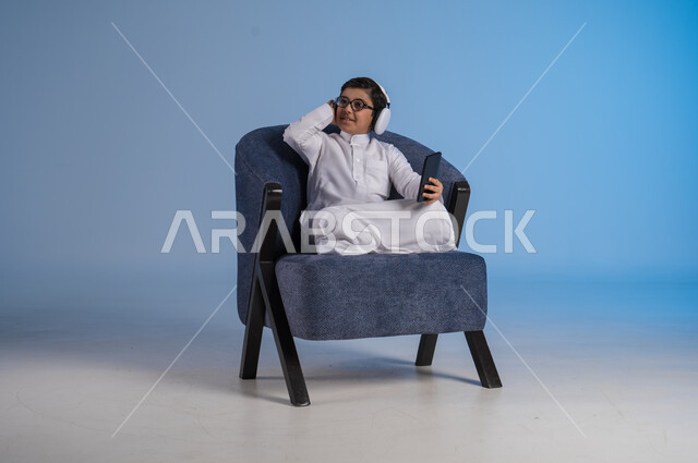 استخدام تقنية الهيدفون الحديثة, قضاء وقت ممتع بالاستماع إلى المقاطع الموسيقية المفضلة, بورتريه لفتى عربي خليجي سعودي يرتدي الثوب التقليدي يجلس على كنبه مريحة يضع سماعة الرأس الصوتية ويستخدم الهاتف المحمول, خلفية زرقاء