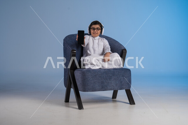 استخدام التقنيات الحديثة المتطورة, بورتريه لولد عربي خليجي سعودي يرتدي الثوب التقليدي وسماعة الهايدفون يقوم بعرض شاشة سوداء فارغة عبر الهاتف المحمول، خلفية زرقاء