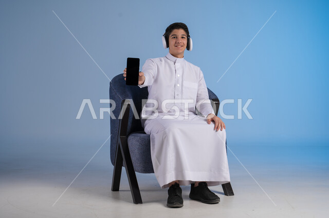 استخدام التقنيات الحديثة المتطورة, بورتريه لشاب عربي خليجي سعودي يرتدي الثوب التقليدي وسماعة الهايدفون يقوم بعرض شاشة سوداء فارغة عبر الهاتف المحمول، خلفية زرقاء