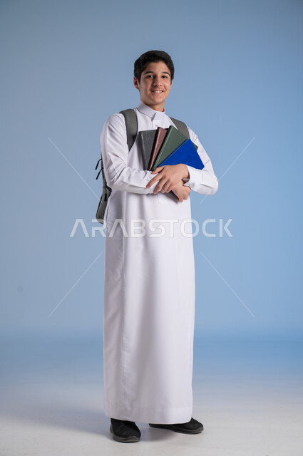 النظر الى الكاميرا بايماءات السرور، مفهوم الاجتهاد للوصول إلى النجاح، المناهج العلمية التعليمية، مفهوم العودة إلى المدارس، بورتريه لطالب عربي خليجي سعودي يرتدي الثوب التقليدي يحمل حقيبة الظهر المدرسية ويحمل مجموعه من الكتب، صورة لكامل الجسم، خلفية زرقاء